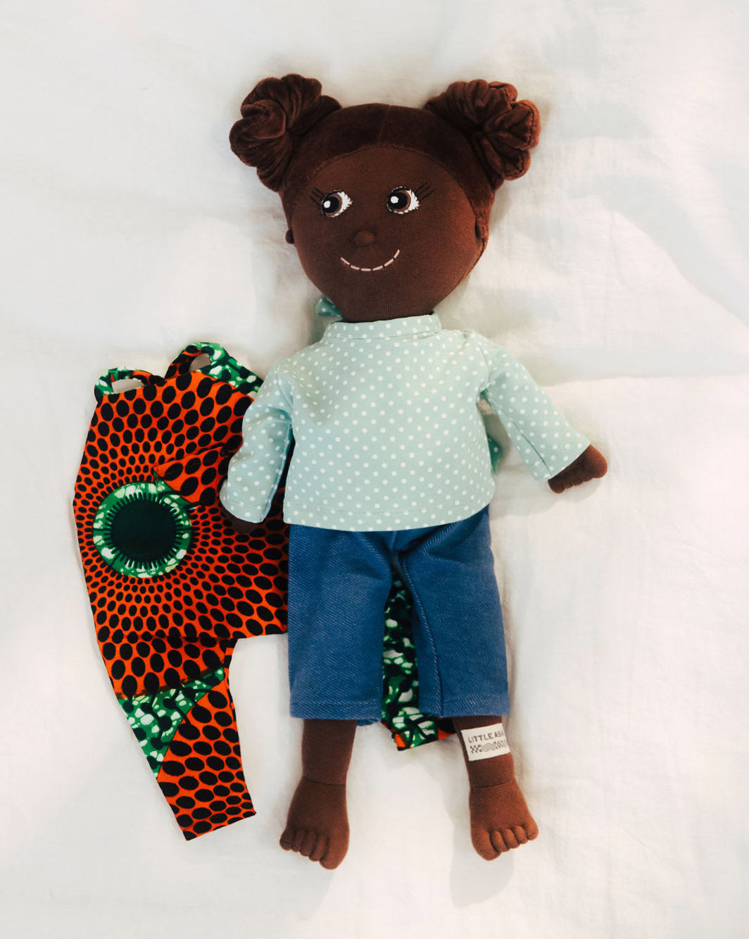 Kinderpuppe mit Haarzöpfen und Puppen Outfit aus Baumwolle. Puppe liegt auf Wickeltisch