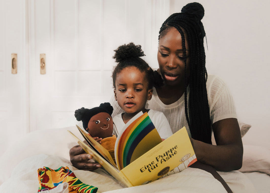 Mutter liest Kinderbuch vor. Kind hält Stoffpuppe in der Hand. Puppenkleidung liegt auf dem weißen Bett