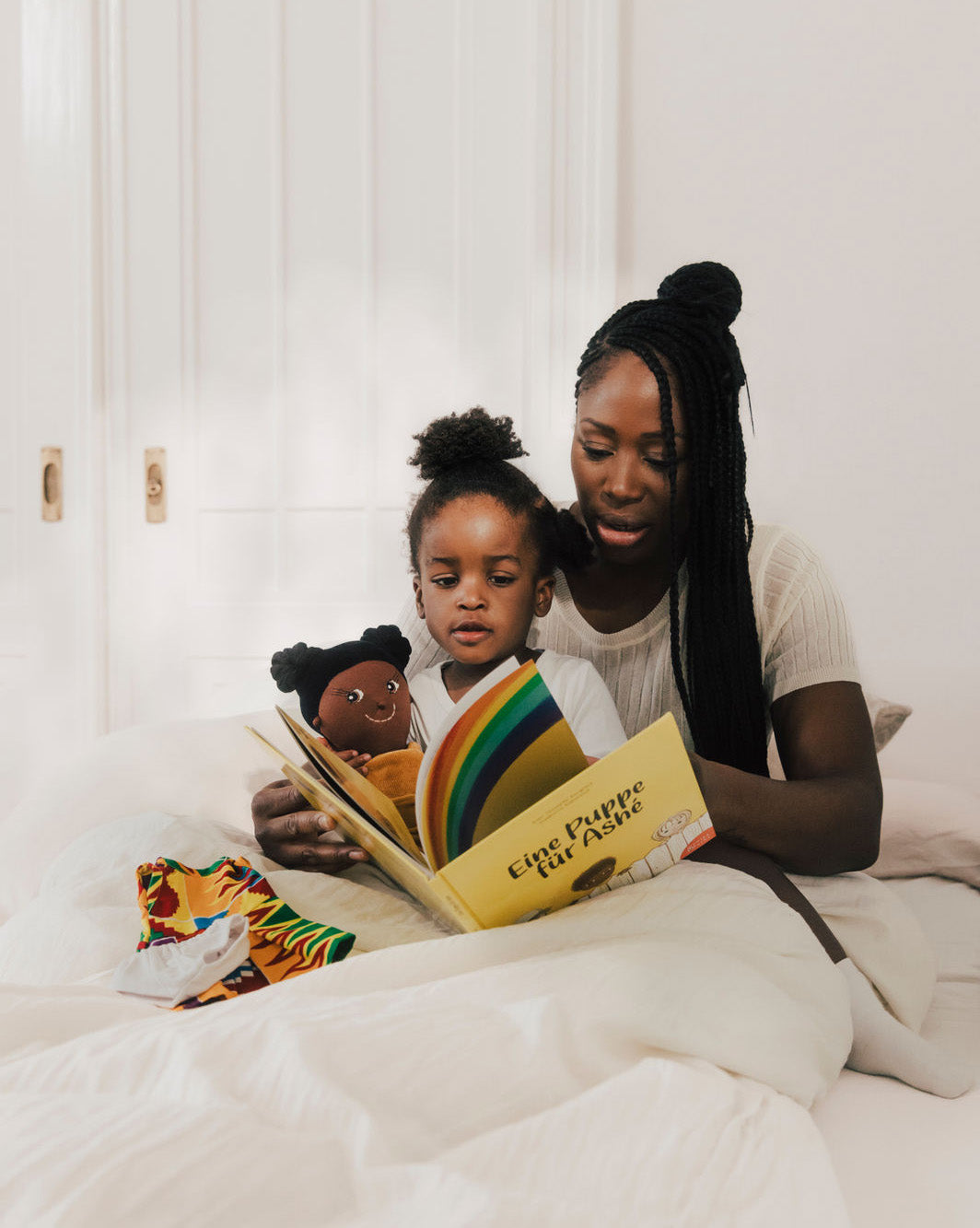 Mutter liest Kinderbuch vor. Kind hält Stoffpuppe in der Hand. Puppenkleidung liegt auf dem weißen Bett
